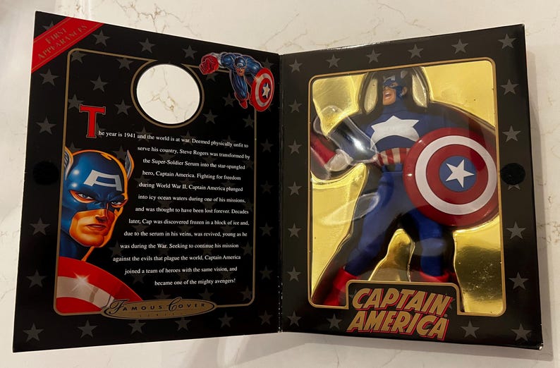 May include: A Captain America action figure in a display box. The figure is in a blue, red, and white costume, holding a shield. The box has a gold interior and black exterior with text that reads "Captain America."