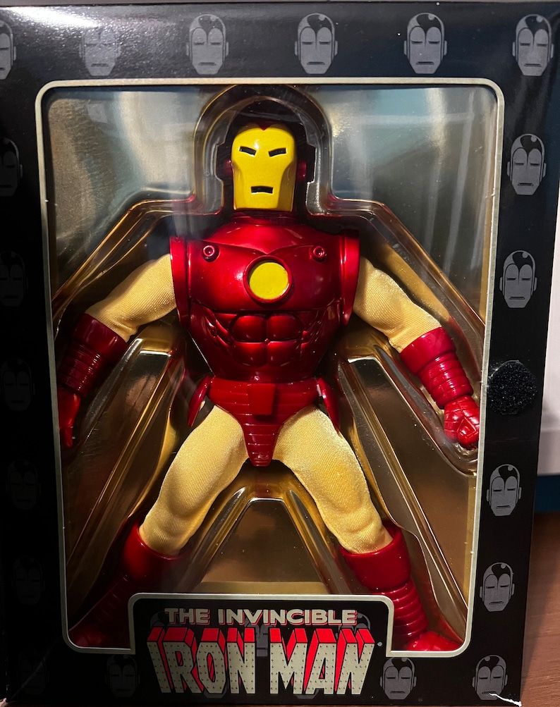 May include: A red and yellow action figure of Iron Man in a clear plastic box. The box has a black background with a repeating pattern of Iron Man's face. The box has the text "The Invincible Iron Man" on the bottom.