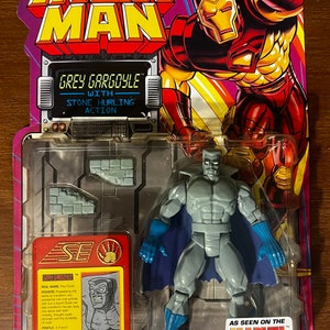 May include: A plastic action figure of Grey Gargoyle from the Marvel Comics universe. The figure is grey with a blue cape and is holding a stone in his right hand. The figure is packaged in a plastic blister pack with a red, white, and blue background. The packaging includes the Marvel logo and the words "As Seen on the Marvel Action Hour".