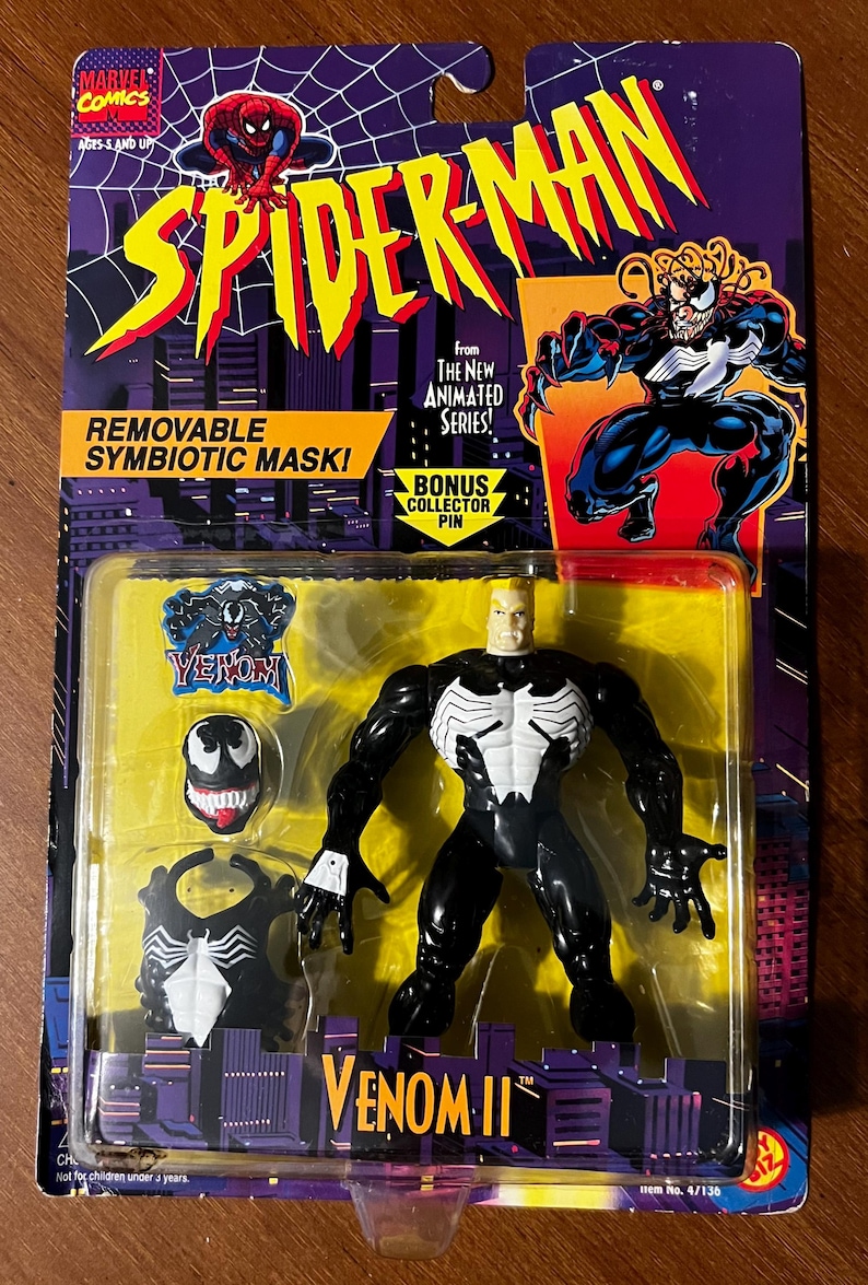May include: A black and white action figure of Venom II from the Spider-Man animated series. The figure has a removable mask and comes with a bonus collector pin. The packaging features a yellow background with the Spider-Man logo and the words "Removable Symbioitic Mask!" and "Bonus Collector Pin".