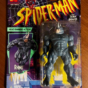 May include: A Spider-Man action figure of Rhino, from the animated series, in its original packaging. The figure is gray and blue, with a horn on its head. The packaging features the Marvel Comics logo, Spider-Man imagery, and the text "HEAD RAMMING ACTION!"