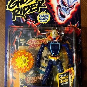 Może przedstawiać: Figurkę akcji Ghost Ridera z elementami świecącymi w ciemności. Figurka ma na sobie niebieski strój z żółtym wzorem płomieni. Figurka jest wyposażona w obracającą się broń łańcuchową i komiks. Opakowanie zawiera tekst "Ghost Rider" i "Eksplodująca akcja tułowia!"