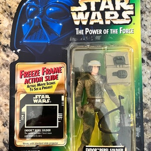 May include: A Star Wars action figure of a Rebel Soldier from the movie Return of the Jedi. The figure is wearing a green camouflage uniform and a helmet. The figure is holding a blaster rifle and has a backpack. The figure is packaged in a plastic blister pack with a green and black background. The packaging includes a freeze frame action slide that shows scenes from the movie. The packaging also includes the text "Star Wars: The Power of the Force".