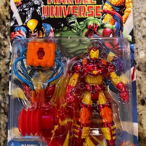 Puede incluir: Una figura de acci&oacute;n de Iron Man de la l&iacute;nea de juguetes Marvel Universe. La figura es roja y dorada y sostiene un accesorio de explosi&oacute;n de energ&iacute;a azul y naranja. El empaque incluye una etiqueta de advertencia sobre los peligros de asfixia.