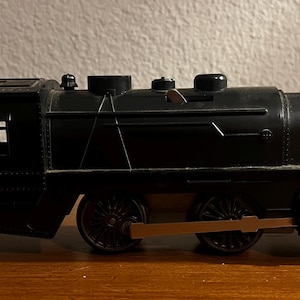 May include: A black toy train with a detailed design. The train features a cab with windows, multiple smokestacks, and visible wheels. The train is made of a dark material and sits on a wooden surface.