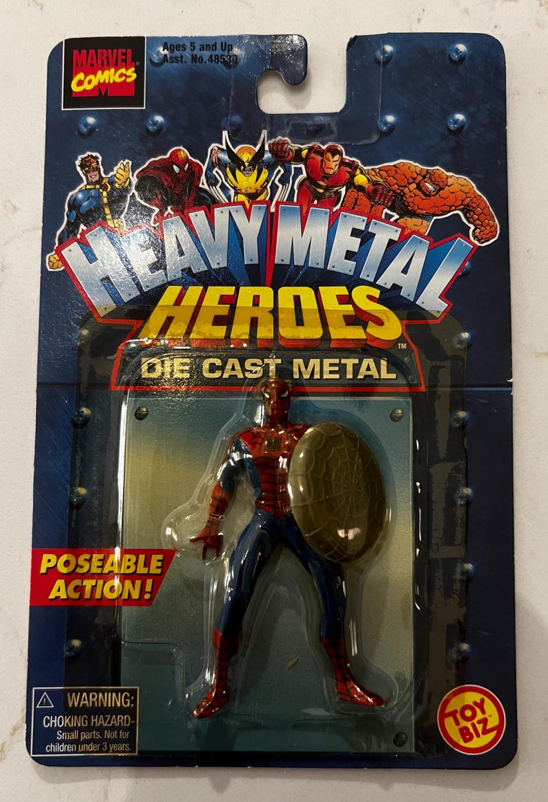 以下が含まれることがあります： マーベルコミックの「ヘビーメタルヒーローズ」スパイダーマンのアクションフィギュア、ダイキャストメタル製。フィギュアは赤と青で、盾を持っています。パッケージにはマーベルコミックのロゴと「Poseable Action！」の文字が含まれています。