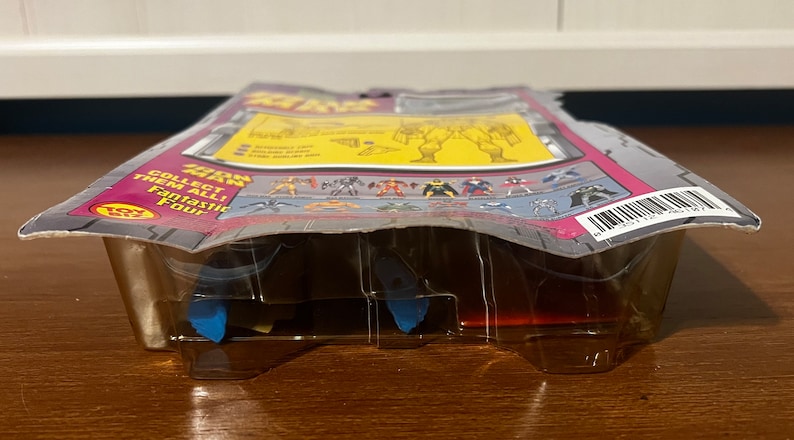 May include: A plastic blister pack containing a Fantastic Four action figure. The packaging is pink, gray, and white with the words "The Thing" and "Collect them all! Fantastic Four" printed on it. The figure is not visible in the packaging.