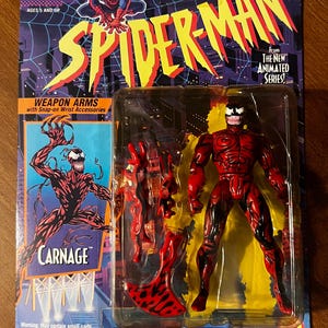 May include: Carnage action figure from the Spider-Man animated series. The figure is red and black with a white spider symbol on the chest. The figure comes with weapon arms and snap-on wrist accessories. The packaging features a cartoon image of Spider-Man and the text "Weapon Arms with Snap-on Wrist Accessories".