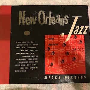 Peut inclure: Une pochette de disque vinyle noire et rouge avec le texte "New Orleans Jazz" dans une police de caractères stylisée. La pochette présente une carte de la Nouvelle-Orléans avec divers lieux et noms de musiciens énumérés sur le côté gauche.