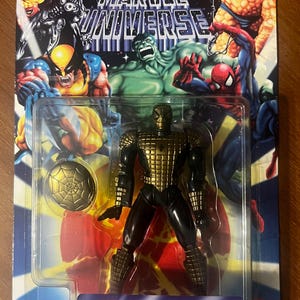 Pode incluir: Uma figura de a&ccedil;&atilde;o dourada e preta do Homem-Aranha com um escudo de teia. A figura &eacute; embalada em uma embalagem blister de pl&aacute;stico com fundo vermelho, amarelo e azul. A embalagem apresenta o logotipo do Marvel Universe e o texto "Launching Web Shield Action".
