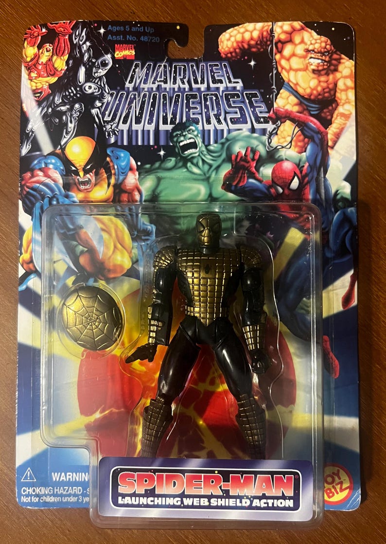 Pode incluir: Uma figura de a&ccedil;&atilde;o dourada e preta do Homem-Aranha com um escudo de teia. A figura &eacute; embalada em uma embalagem blister de pl&aacute;stico com fundo vermelho, amarelo e azul. A embalagem apresenta o logotipo do Marvel Universe e o texto "Launching Web Shield Action".