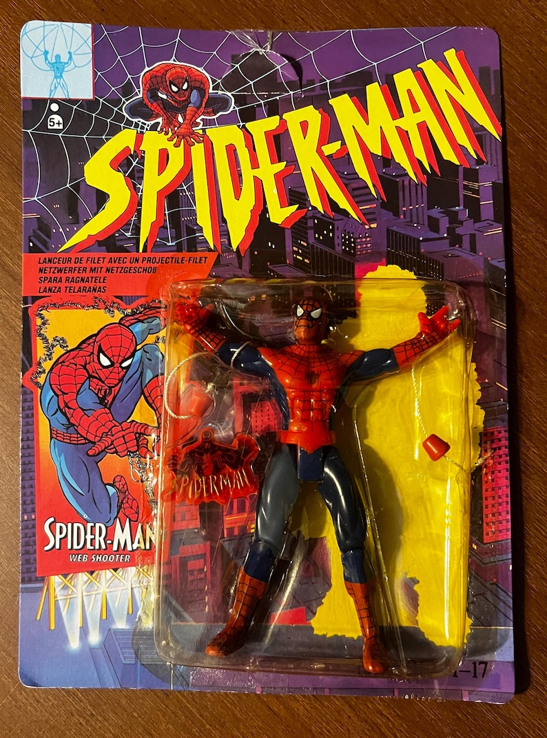 May include: A red and blue Spider-Man action figure with a web shooter accessory. The figure is packaged on a yellow and purple card with the text "SPIDER-MAN" in large yellow letters.