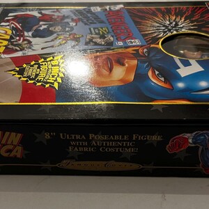 May include: A black box featuring Captain America artwork. The box includes the text "Captain America" and "8 inch Ultra Poseable Figure with Authentic Fabric Costume!" The box has a clear window showing the figure.