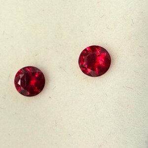 May include: Two round, faceted, deep red gemstones. Each stone features a brilliant cut with multiple triangular facets that reflect light, creating a vibrant, saturated crimson color throughout the circular surface of the gems.