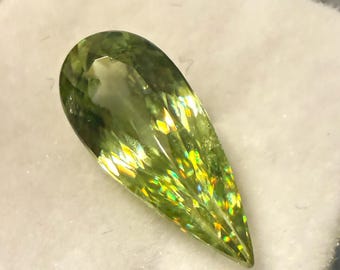 May include: A pear-shaped, faceted gemstone with a light green hue. The gem has internal rainbow-like flashes of color and is set against a white background. The gemstone is likely for jewelry making.