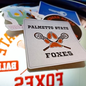 May include: A square sticker featuring the text PALMETTO STATE at the top and FOXES at the bottom. In the center, two crossed lacrosse sticks are positioned behind an orange paw print graphic. The sticker is white with dark brown lettering.
