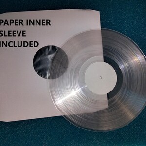 K&ouml;nnte beinhalten: Eine transparente Vinyl-Schallplatte mit wei&szlig;em Label, neben einer wei&szlig;en Papier-Innenh&uuml;lle. Auf der H&uuml;lle steht der Text "PAPER INNER SLEEVE INCLUDED". Die transparente Schallplatte l&auml;sst Licht durchscheinen.