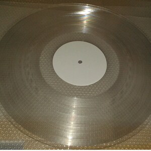 K&ouml;nnte beinhalten: Eine transparente Schallplatte mit einem wei&szlig;en Label in der Mitte. Die Platte ist klar und zeigt konzentrische Rillen. Das Label hat ein kleines Loch in der Mitte.