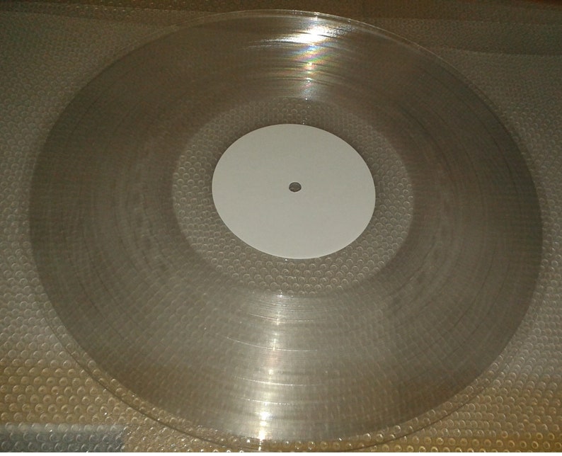 K&ouml;nnte beinhalten: Eine transparente Schallplatte mit einem wei&szlig;en Label in der Mitte. Die Platte ist klar und zeigt konzentrische Rillen. Das Label hat ein kleines Loch in der Mitte.