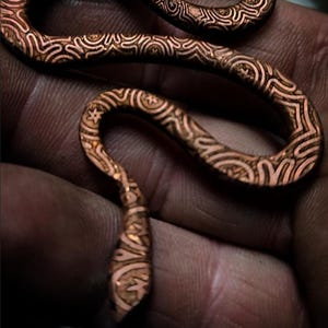 May include: A copper-colored, intricately patterned snake sculpture. The snake is coiled and detailed with a repeating design. The sculpture is held in a hand, showcasing the craftsmanship and texture of the metalwork.