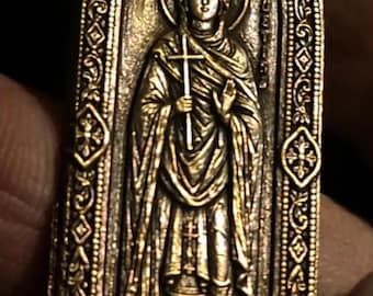May include: An oval-shaped bronze-toned metal pendant featuring a relief of a robed figure holding a cross. The border is decorated with intricate filigree patterns and geometric motifs. A small metal loop is attached to the top for suspension.