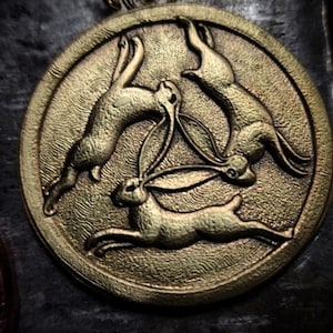 May include: A gold-toned metal pendant featuring three stylized rabbits in a circle, forming a triquetra design.