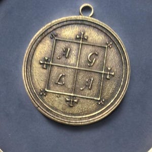May include: A brass pendant with a square design featuring the letters "I", "G", "L", and "I" in a grid pattern. The pendant is surrounded by a circular frame with decorative details.