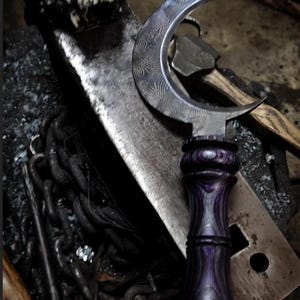 Puede incluir: Un cuchillo en forma de media luna con un mango de madera púrpura. El cuchillo está apoyado en una superficie de metal con una calavera en el fondo.
