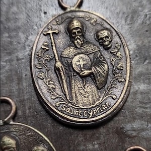 May include: A gold-toned metal pendant featuring a relief image of Saint Cyprian, a Christian saint, holding a cross and a skull. The pendant is oval-shaped and has a loop at the top for hanging.
