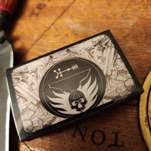 May include: A black and white playing card with a skull and wings design. The card has a circle with a symbol and the text "TETRA" and "NOL" on the background.