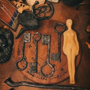 May include: A collection of mystical items on a wooden surface. Includes three ornate metal keys, a candle figure, and other ritualistic objects. The keys are arranged on a circular wooden board with symbols.