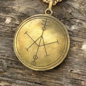 May include: A gold-toned pendant necklace with a circular brass charm featuring a geometric design. The charm has a raised edge and a textured surface.