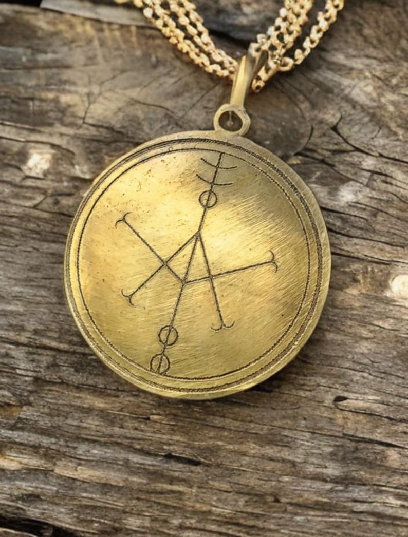 May include: A gold-toned pendant necklace with a circular brass charm featuring a geometric design. The charm has a raised edge and a textured surface.