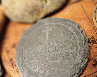 May include: A grey slate coaster featuring a decorative design of a cross and a circle. The coaster is on a wooden surface with a patterned design.
