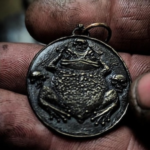 May include: A bronze pendant featuring a detailed frog with skulls on either side. The pendant is round and has a textured surface.