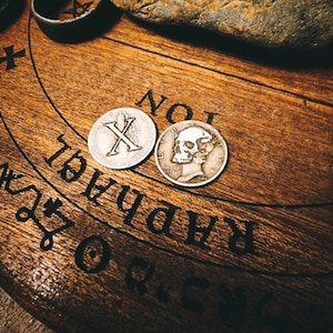 May include: Two silver coins on a wooden board with a Ouija board design. One coin has a skull and crossbones, the other has a Roman numeral X.
