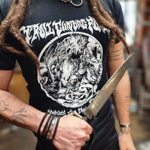 May include: A black t-shirt with a white graphic print of a creature with tentacles and a skull. The text "Troll Tongue Fury" is printed above the graphic. The person wearing the shirt is holding a large, rusty knife.