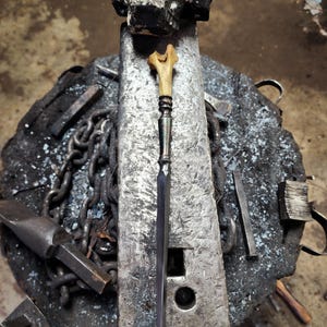 May include: A long, thin, black and silver knife with a bone handle. The knife is resting on a metal anvil with a skull on top.