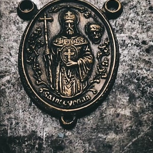 May include: A detailed, antique-style bronze medallion featuring a religious figure holding a cross and staff. The inscription "Saint Cyprian" is visible. The medallion has a textured, aged appearance, with a dark background and decorative elements.