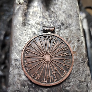 May include: A copper-colored pendant with a circular design featuring a sunburst pattern and various symbols around the edge. The pendant has a loop at the top for attaching a chain.