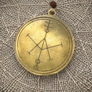 May include: A brass pendant with a circular design featuring a geometric symbol. The pendant has a loop at the top for hanging.
