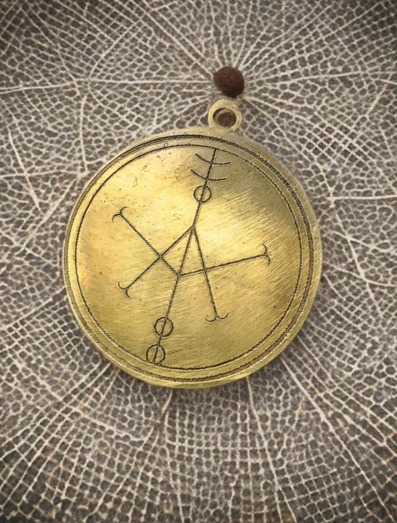 May include: A brass pendant with a circular design featuring a geometric symbol. The pendant has a loop at the top for hanging.