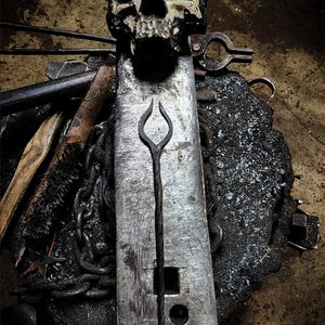 May include: A blacksmith's workspace featuring a skull atop an anvil. A long, slender, metal tool with a looped end is centered on the anvil. Various tools, including tongs and chains, surround the anvil, creating a dark, industrial aesthetic.