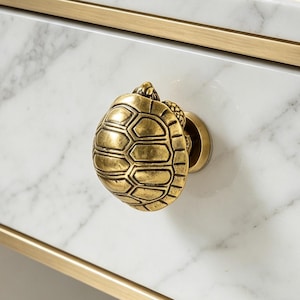 May include: A brass turtle-shaped drawer knob mounted on a white marble drawer front with grey veining. The drawer is framed in a brushed gold metal structure, featuring a clean, rectangular design with a smooth, polished stone surface finish.