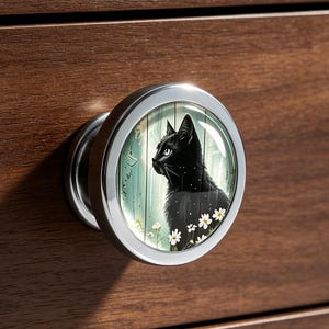 May include: A round silver cabinet knob featuring a glossy insert with a black cat illustration. The cat is depicted in profile against a light green background with small white flowers. The knob is mounted on a dark brown wooden drawer front.