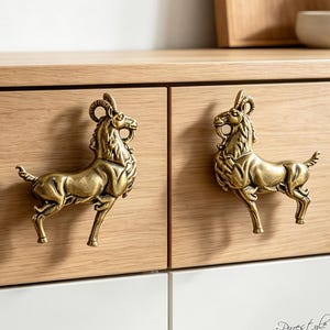 Puede incluir: Dos pomos de armario de metal color latón con forma de cabras de pie, con cuernos curvados y texturas de pelaje detalladas. Los pomos están montados en un frente de cajón de madera clara y presentan un acabado metálico con reflejos oscuros.