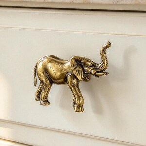 May include: A brass-toned metal drawer pull shaped like a standing elephant with its trunk raised, mounted on a cream-coloured wooden drawer front. The elephant figure features detailed textures and a metallic finish, positioned centrally on the drawer.