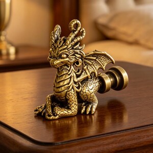 Pode incluir: Um remate de lâmpada de metal em tom de latão com a forma de um pequeno dragão com escamas, asas e chifres detalhados. O dragão está numa posição sentada com uma base cilíndrica roscada que se estende das costas para fixação na lâmpada.