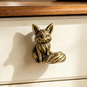 Könnte beinhalten: Ein messingfarbener Metall-Schubladenknopf in Form eines sitzenden Fuchses mit detaillierter Fellstruktur und spitzen Ohren. Der Fuchs ist in sitzender Position dargestellt, wobei sein Schwanz seitlich eingerollt ist, montiert an einer cremefarbenen Schubladenfront.