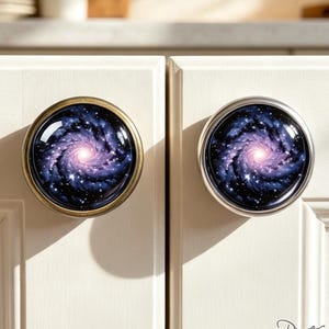 May include: Two round cabinet knobs attached to white cupboard doors. Each knob features a circular insert displaying a deep space galaxy pattern with swirling purple and blue nebulae, bright white stars, and a glowing light center.